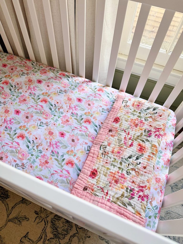 A white crib with a floral-patterned sheet and a matching floral quilt, positioned near a window with natural light coming in.