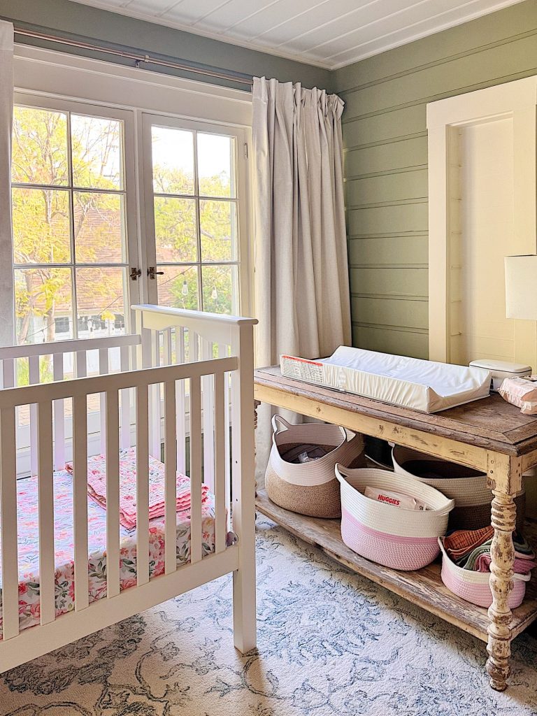 A nursery room with a white crib, a rustic wooden changing table with storage baskets, and large windows with curtains letting in natural light.