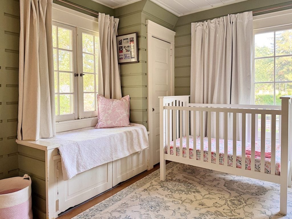 A cozy nursery room with a white crib, a window seat with a pink pillow, beige curtains, green paneled walls, and a light patterned rug.