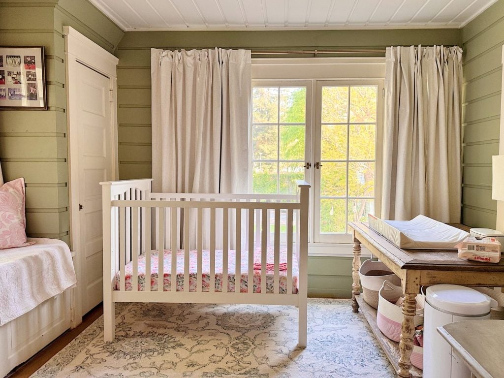 A nursery with a white crib, changing table with supplies, a bench with pink pillows, light curtains, and a large window letting in natural light.