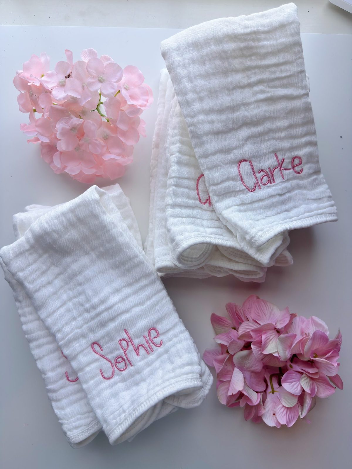 Three white cloths with pink embroidered names "Sophie" and "Clarke" are arranged with two bunches of light pink hydrangea flowers on a white surface.