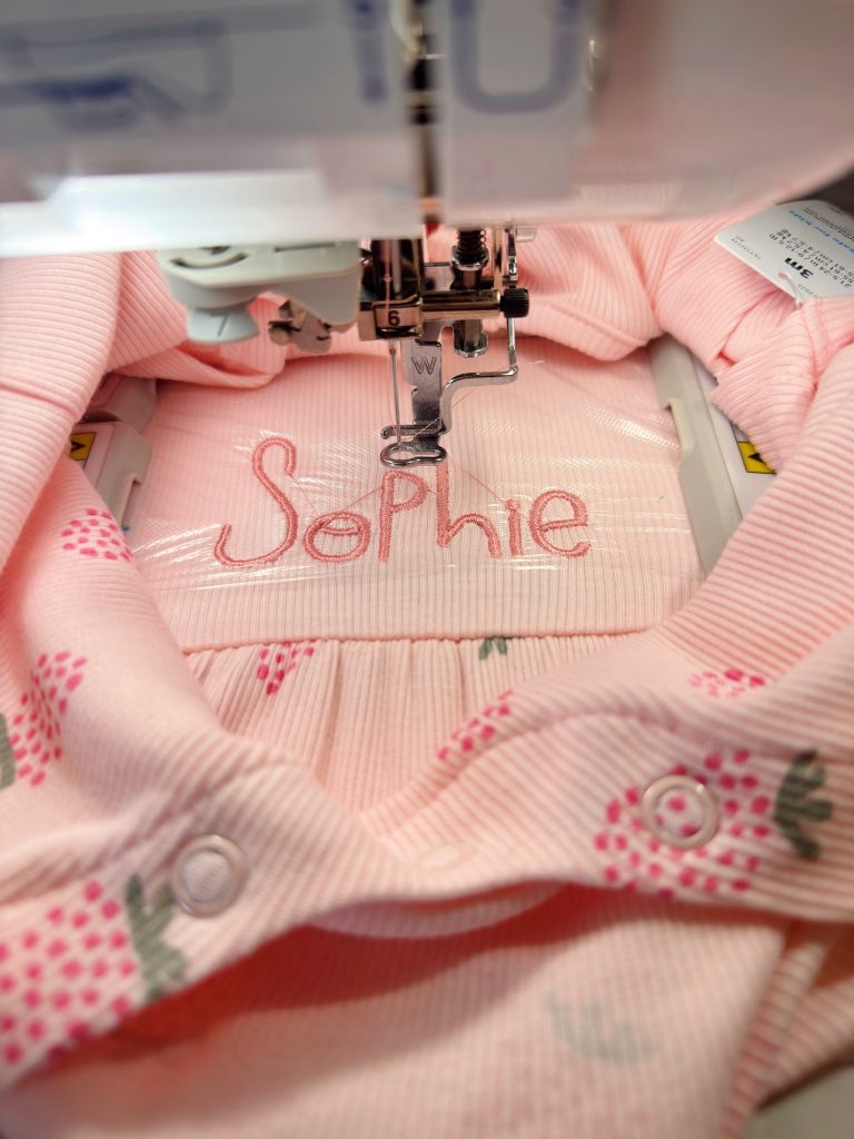 A sewing machine embroiders the name "Sophie" in pink thread on a light pink fabric with a strawberry pattern.