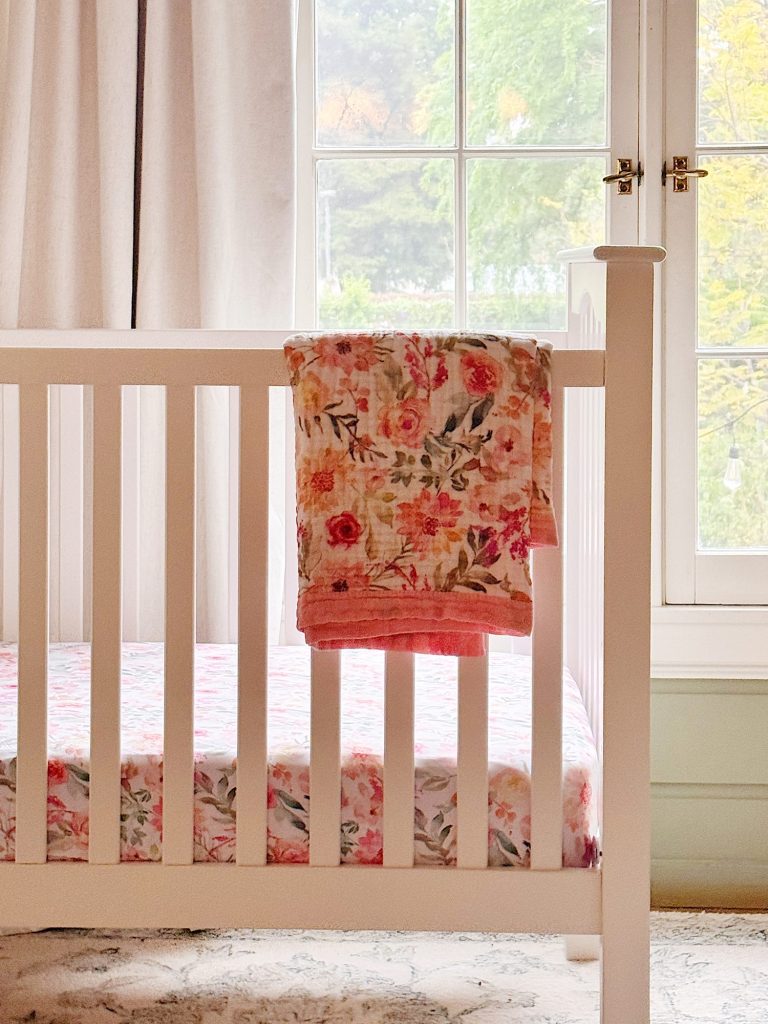 A white wooden crib with a floral blanket draped over the side, positioned in front of a large window with curtains.