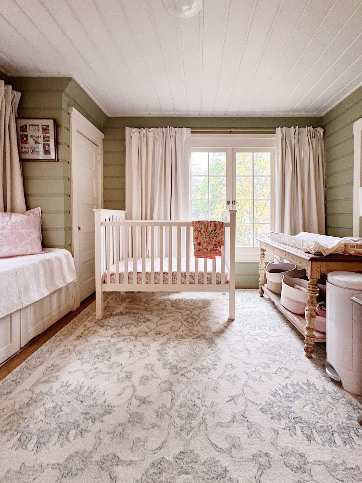 A nursery room with a white crib, a changing table, a bed, light-colored curtains, and a patterned rug on the floor.