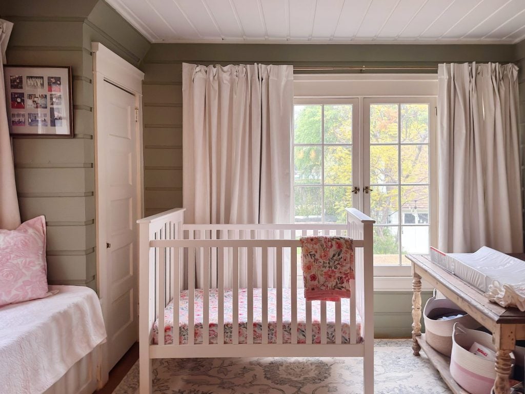 A neatly arranged nursery features a white crib, changing table with baskets, a daybed, light curtains, and a large window overlooking trees.