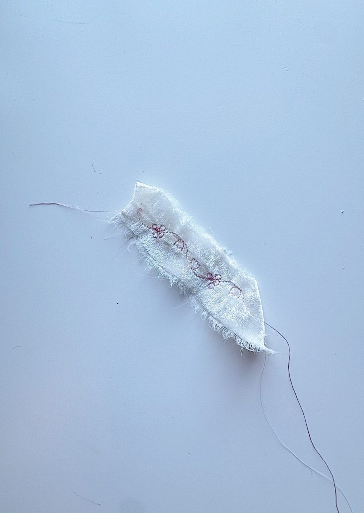 A small piece of frayed white fabric with a line of pink zigzag stitching and loose purple thread on a light surface.