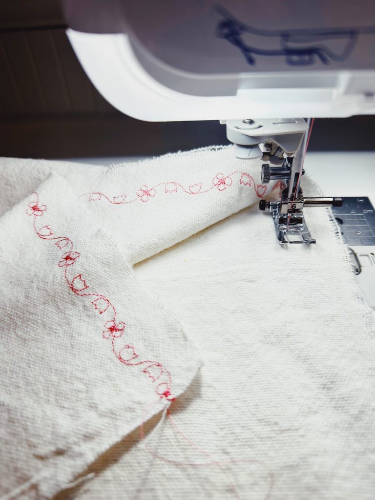 A close-up of a sewing machine stitching red decorative patterns on white fabric.