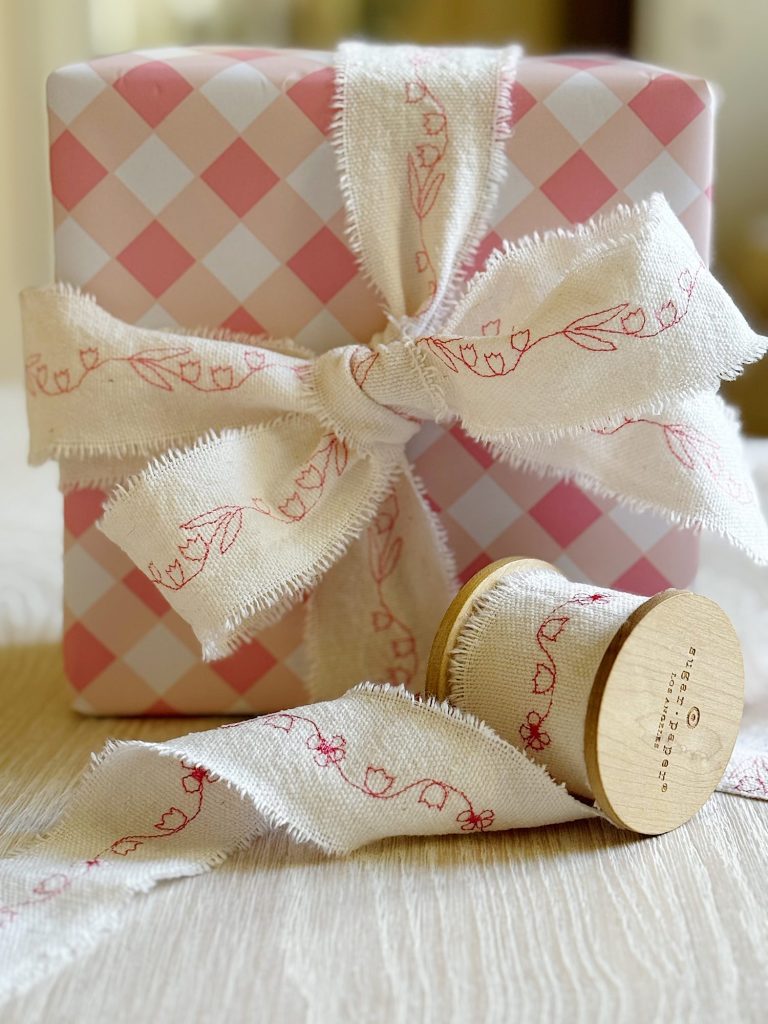 A gift wrapped in pink and white checkered paper with a frayed white fabric ribbon decorated with red floral designs, next to a matching spool of ribbon.