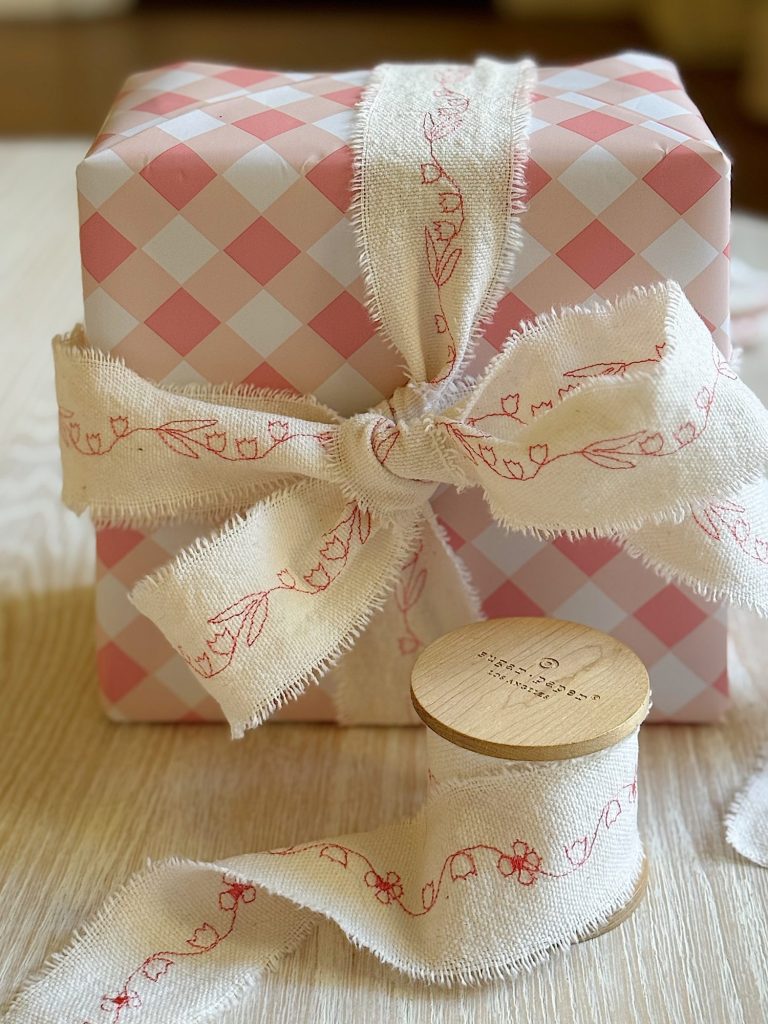 A pink and white checkered gift box wrapped with a frayed ivory ribbon featuring red floral designs, with a matching ribbon spool nearby on a light wood surface.