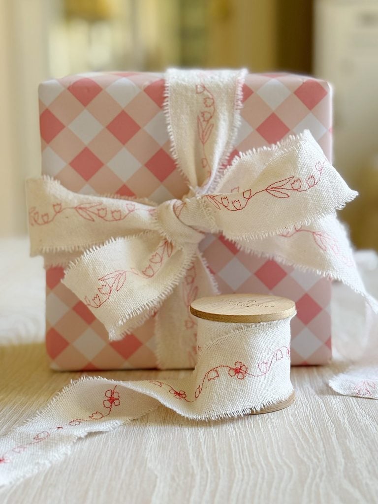 A pink and white checkered gift box tied with frayed white ribbon featuring red floral embroidery, next to a wooden spool of the same ribbon on a light wooden surface.