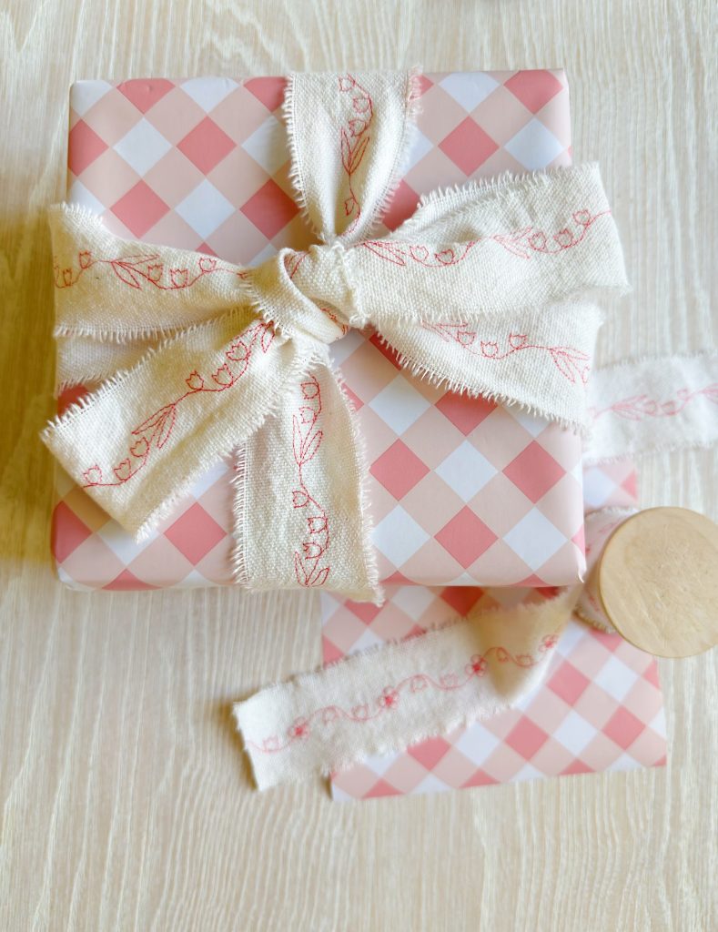 A gift box wrapped in pink and white checkered paper with a frayed white ribbon and floral pattern, placed on a light wooden surface.