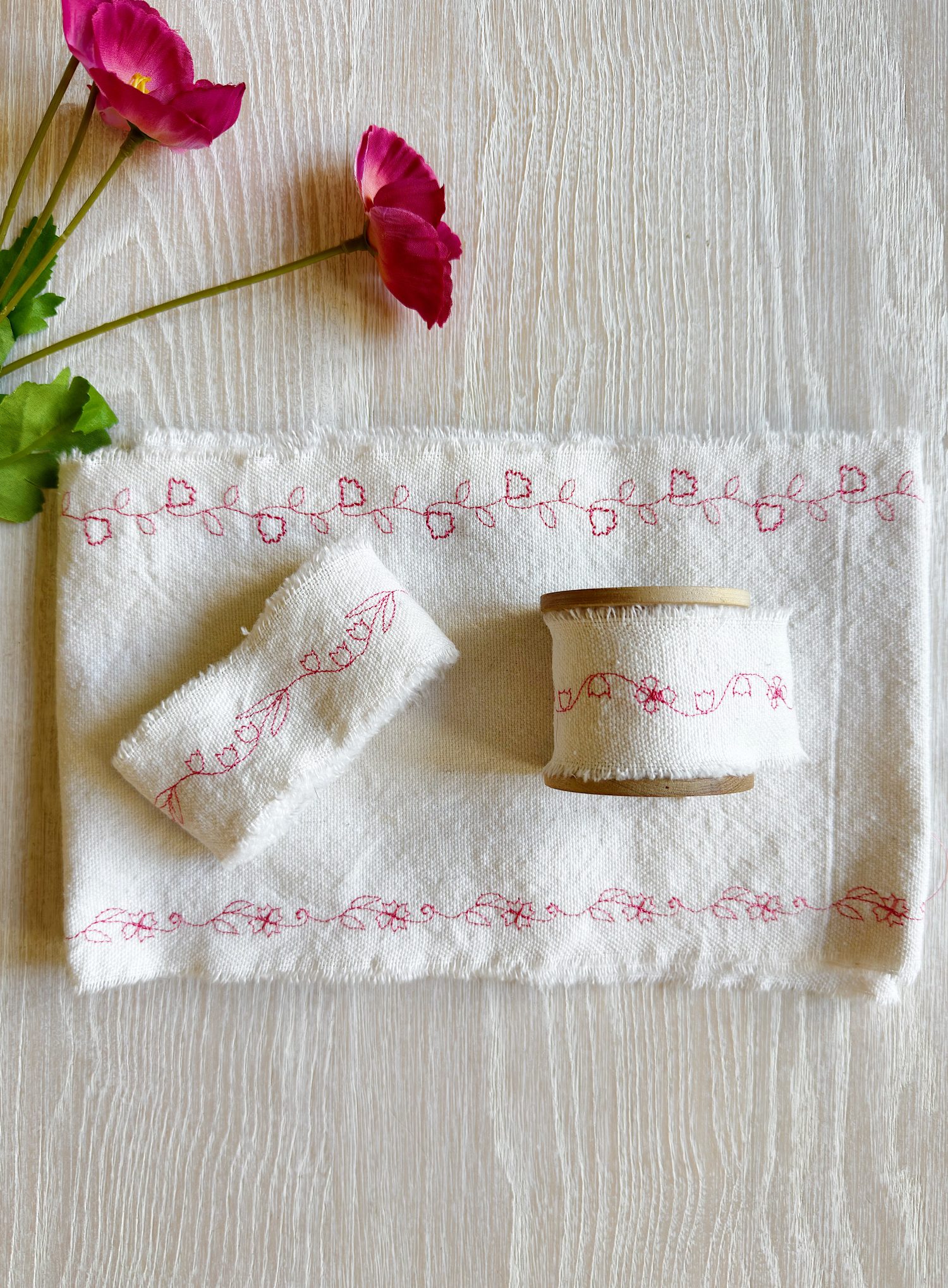 A spool and a roll of white fabric ribbon with pink floral embroidery are displayed on a matching cloth, next to two pink flowers, on a textured white surface.