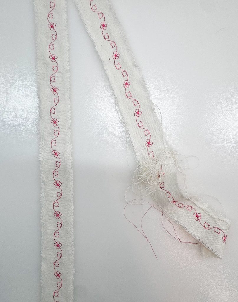 Two strips of white fabric with frayed edges, decorated with a red floral embroidery pattern, are lying on a white surface. One strip has tangled threads at the end.