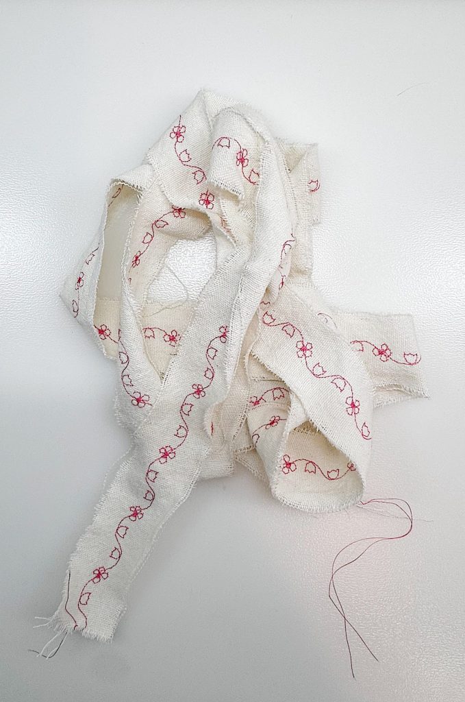 A strip of white fabric with red floral embroidery lies loosely coiled on a white surface, with a pink thread extending from one end.