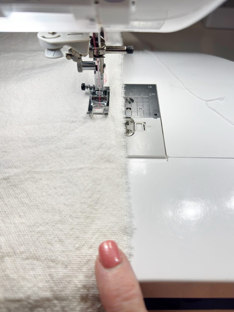 A close-up of a sewing machine stitching a seam in light-colored fabric, with a person's hand guiding the material.