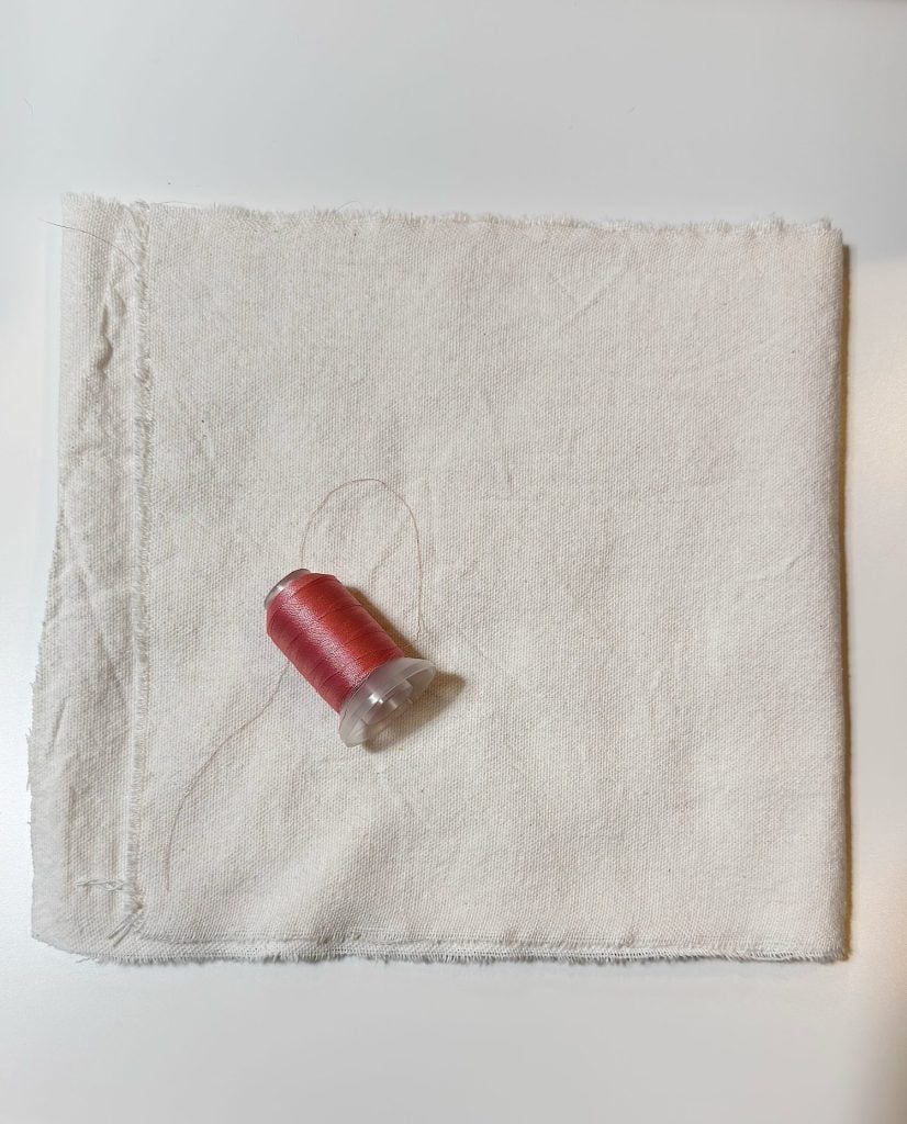 A spool of red thread rests on a folded piece of off-white fabric, placed on a smooth white surface.