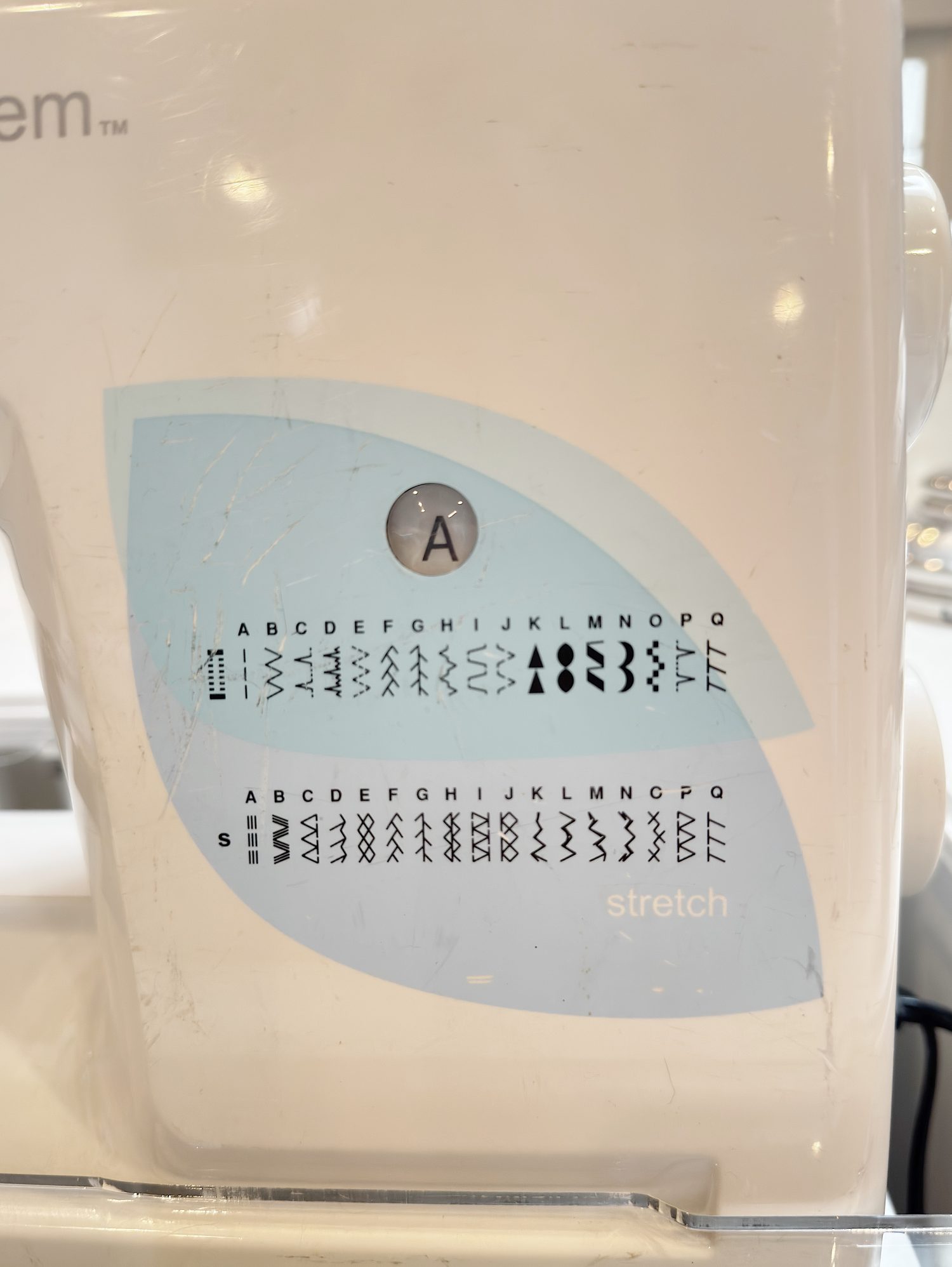 Close-up of a sewing machine stitch selection panel showing various letter-coded stitch patterns, including straight, zigzag, and stretch stitches.
