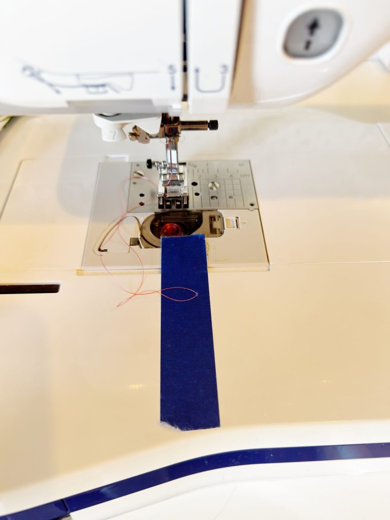 A close-up of a sewing machine with a piece of blue tape placed on the needle plate, guiding the fabric alignment.