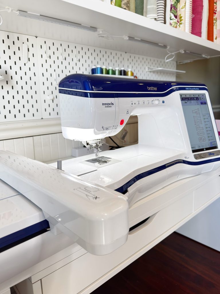 A computerized Brother sewing and embroidery machine sits on a white table, with shelves of thread spools and fabric in the background.