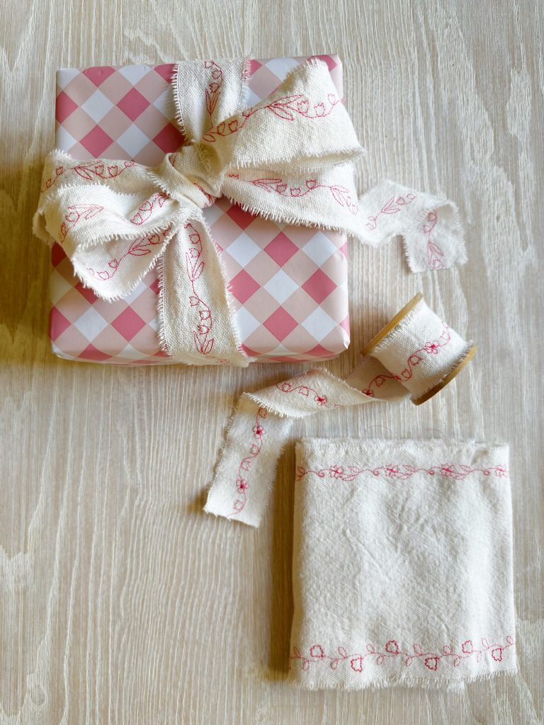 A gift wrapped in pink and white checkered paper with a white fabric ribbon, a matching folded cloth, and a spool of ribbon on a light wood surface.