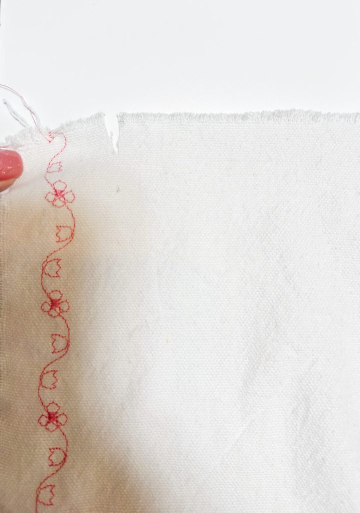 A hand holds a piece of white fabric with a red decorative floral stitch running along the left edge.