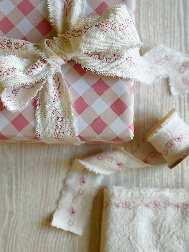 A gift wrapped in pink and white checkered paper with a frayed white ribbon featuring red embroidery, alongside a spool of the same ribbon on a light textured surface.
