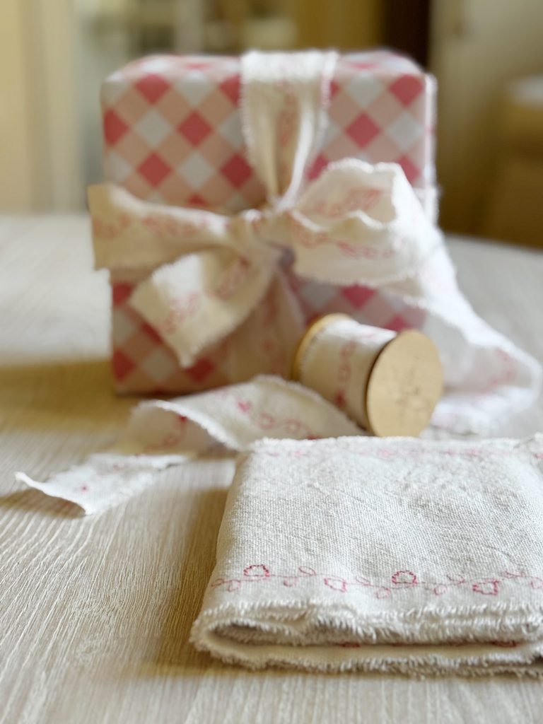 A neatly wrapped gift with a pink and white checkered pattern, topped with a fabric bow, sits on a wooden surface next to a spool of ribbon and a folded cloth.