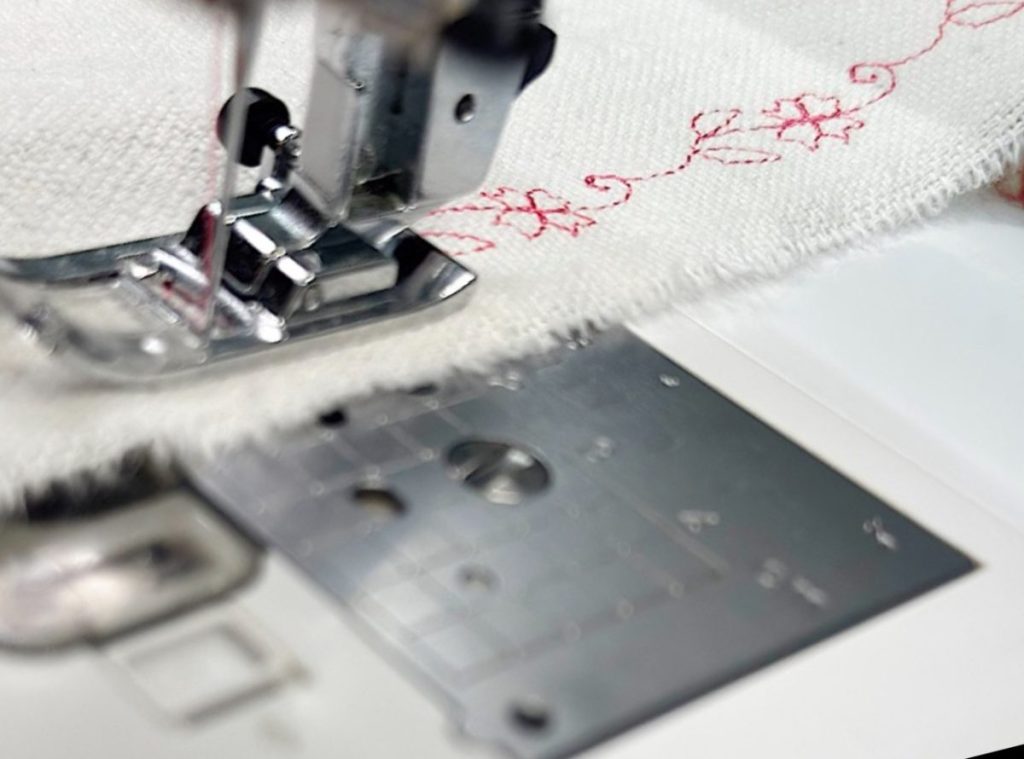 Close-up of a sewing machine stitching red floral patterns on a piece of white fabric.