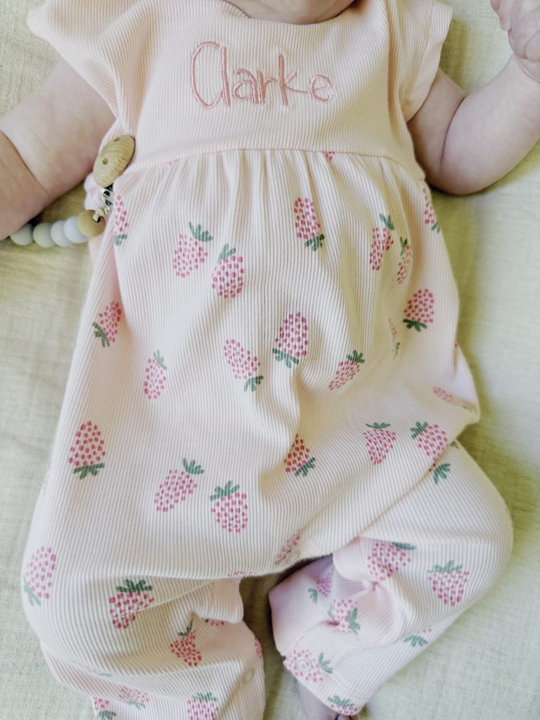 A baby wearing a light pink outfit with a strawberry print and the name "Clarke" embroidered on the chest.