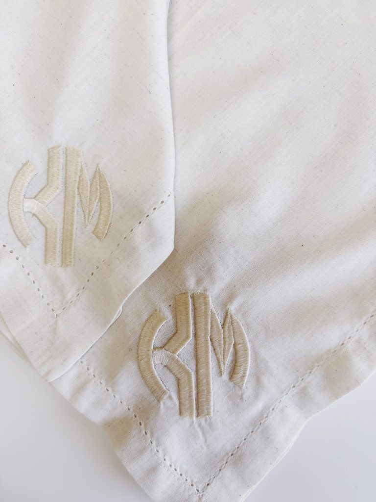 Two white cloth napkins featuring identical beige embroidered monograms with the letters "KM" and visible corner stitching.
