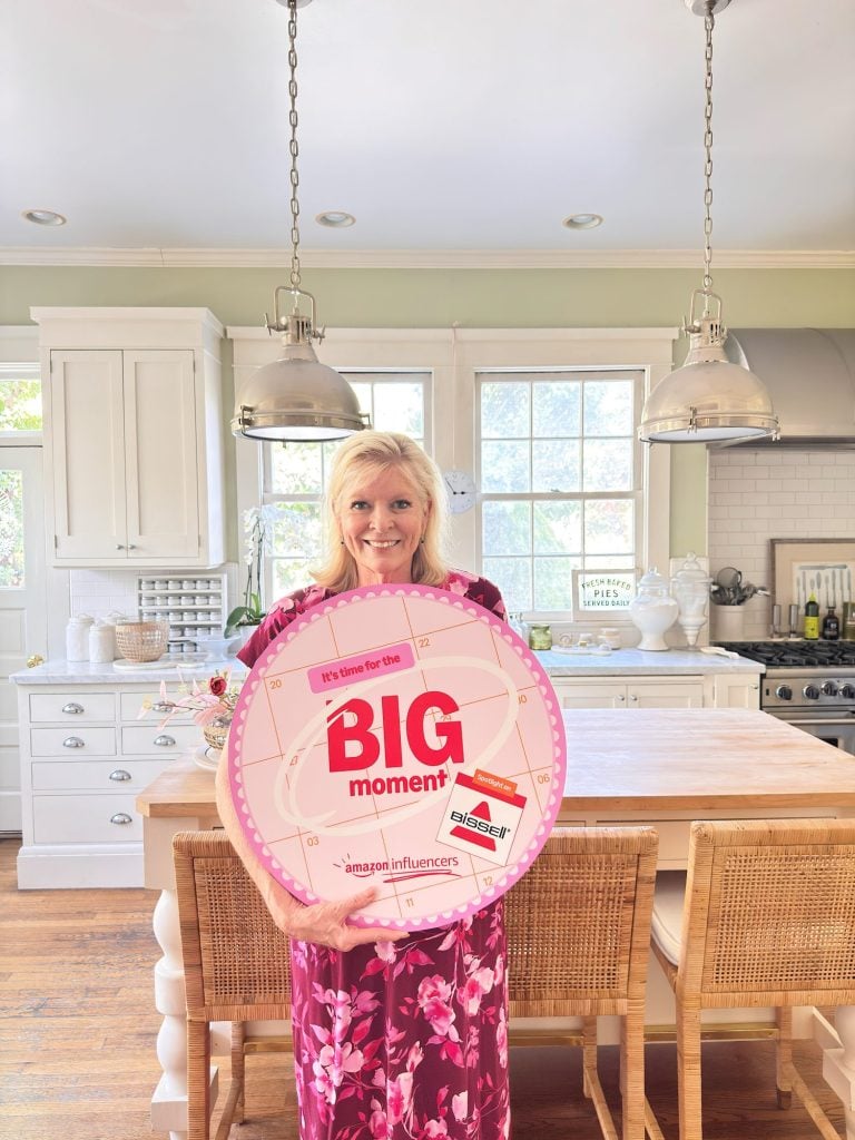 Woman in a pink floral dress stands in a bright kitchen holding a large round sign that says "BIG moment" with various sponsor logos.