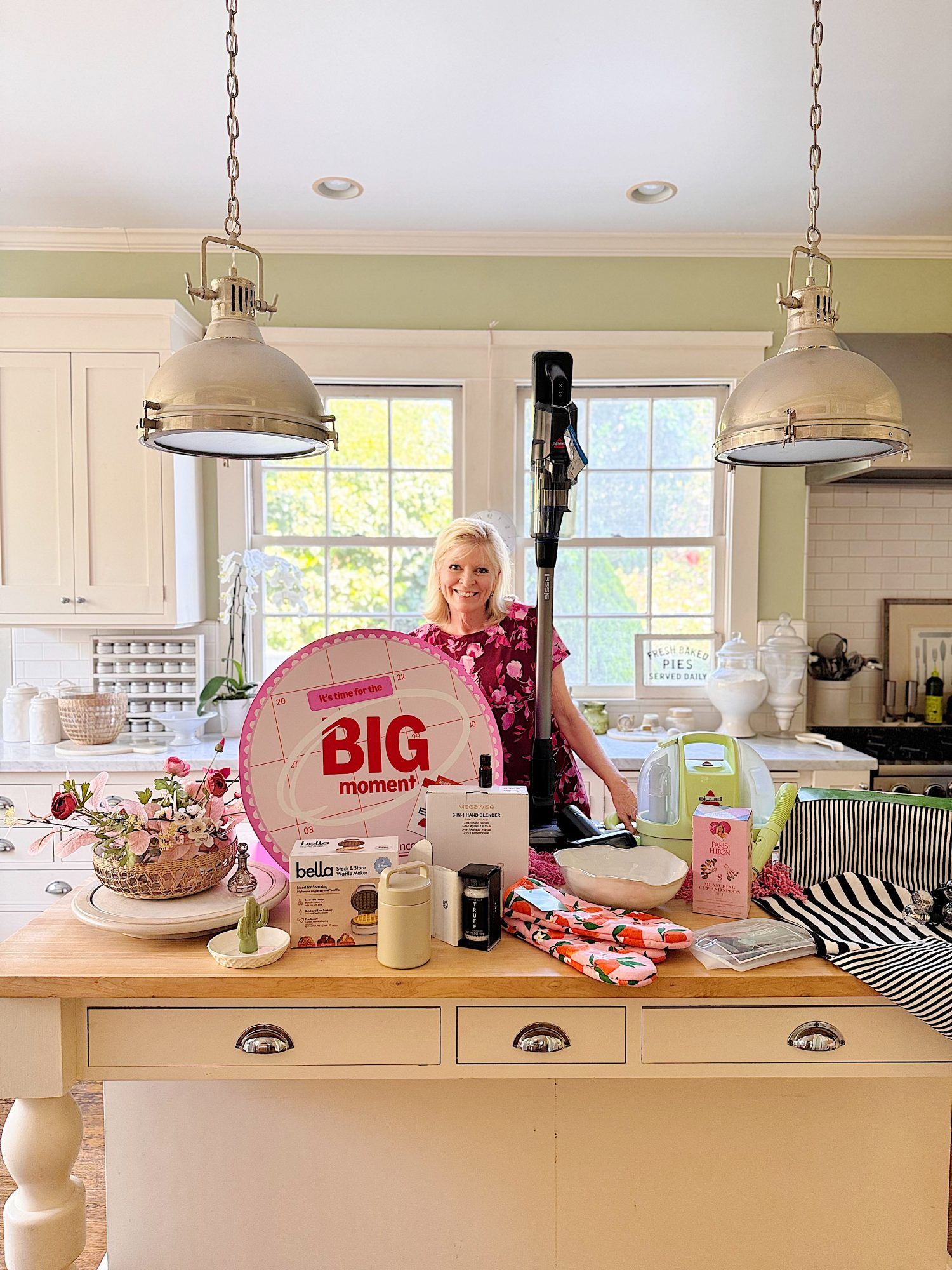 A woman stands in a bright kitchen behind a counter displaying assorted household items, including a large pink "Big Moment" wheel, electronics, kitchenware, and textiles.