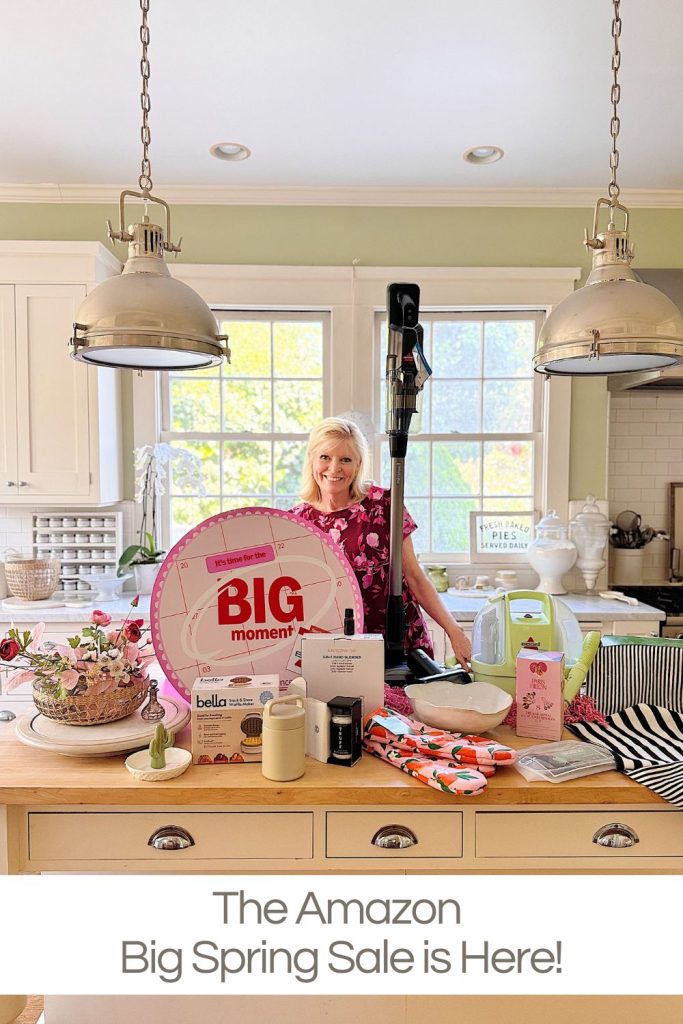 A woman stands in a bright kitchen behind a counter displaying various products with a sign reading "BIG moment" and text below: "The Amazon Big Spring Sale is Here!.