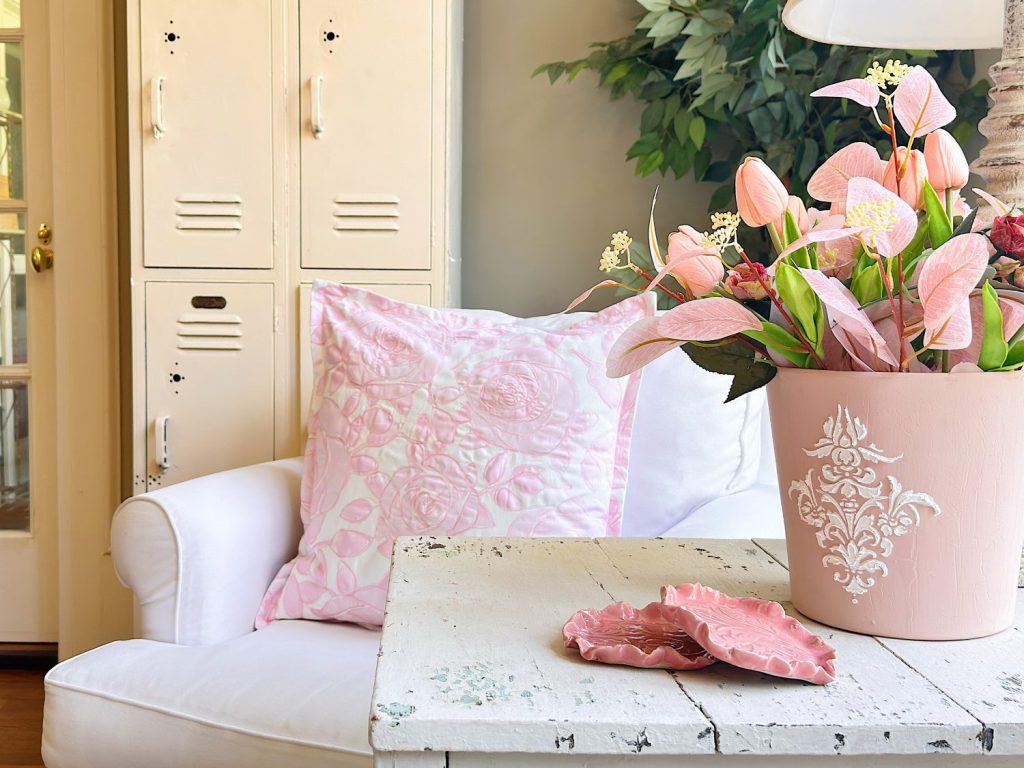 A white armchair with a pink patterned pillow sits beside a white distressed table holding a pink flower pot with artificial flowers, next to cream lockers and a green plant.