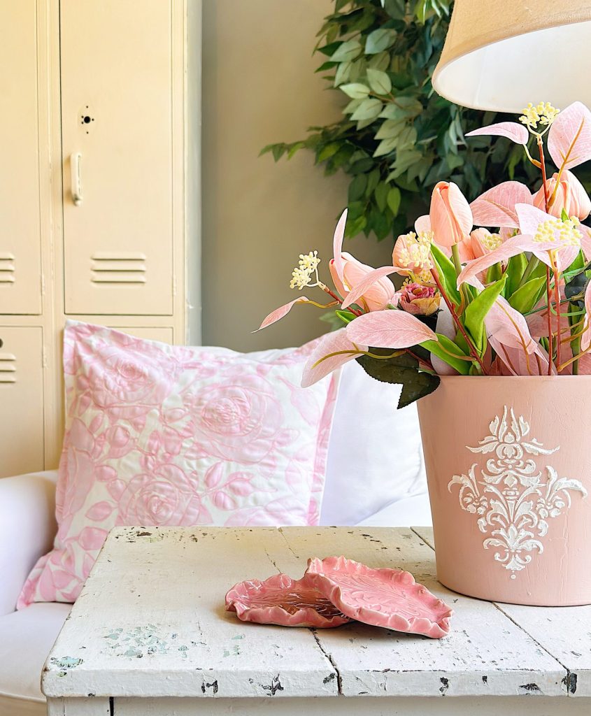 A white chair with a pink-patterned pillow is next to a distressed white table holding pink ceramic dishes and a pink pot with artificial pink and white flowers under a lamp.
