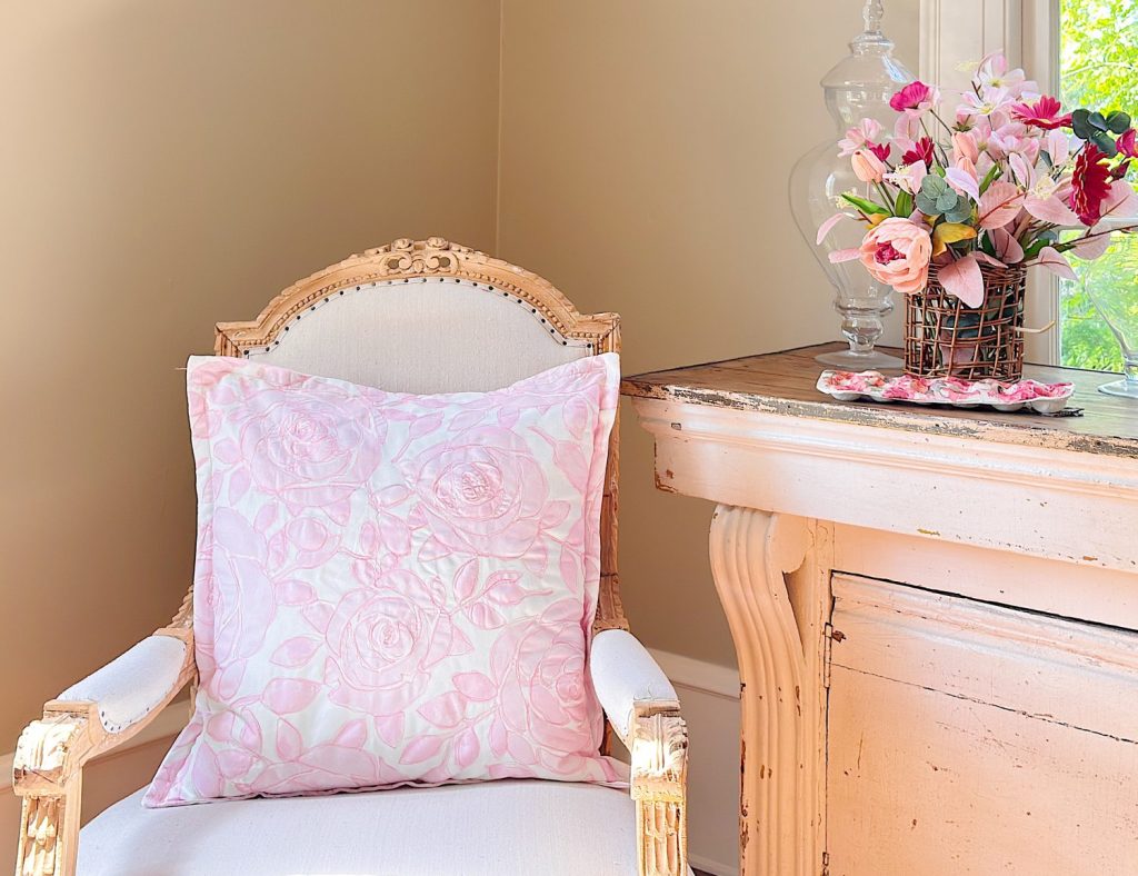 A cream-colored armchair with a pink floral cushion sits beside a distressed white table topped with a vase of pink flowers, near a sunlit window.