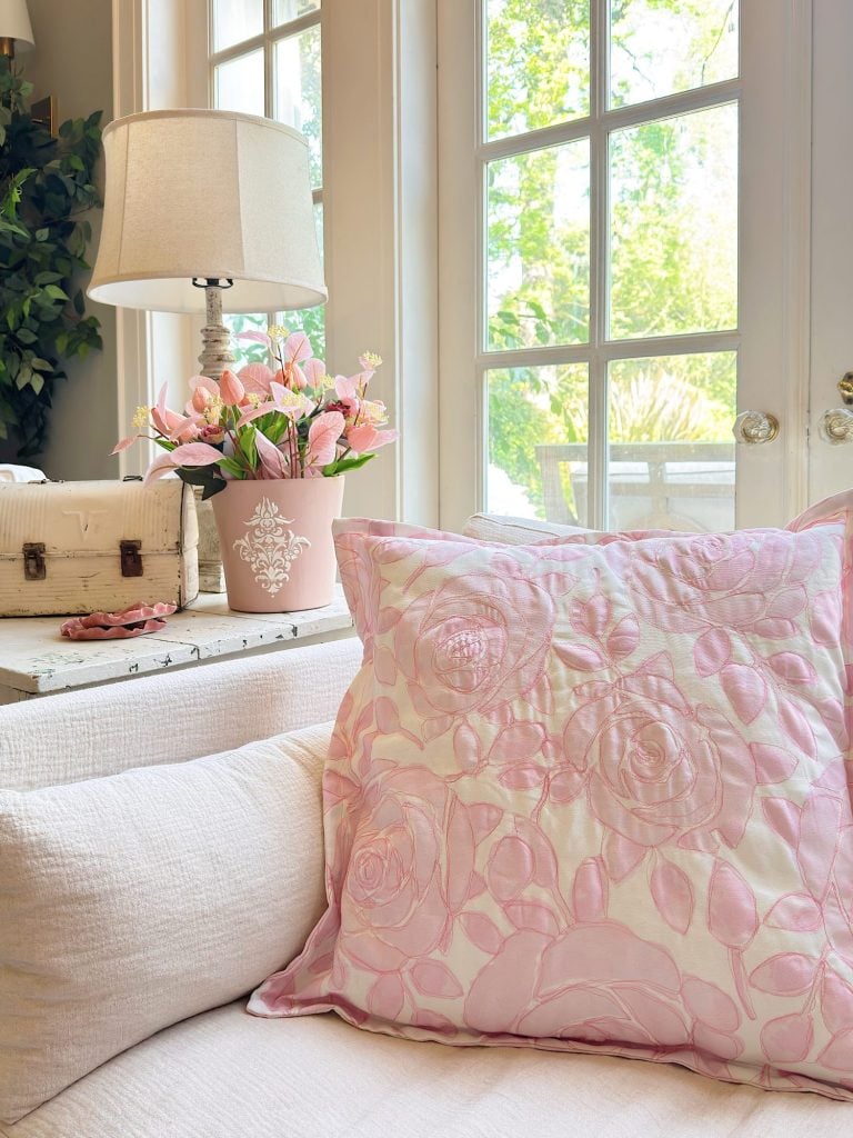 A pink floral pillow on a white couch, with a pink flower arrangement, a white lamp, and a decorative box on a table in front of large windows letting in daylight.