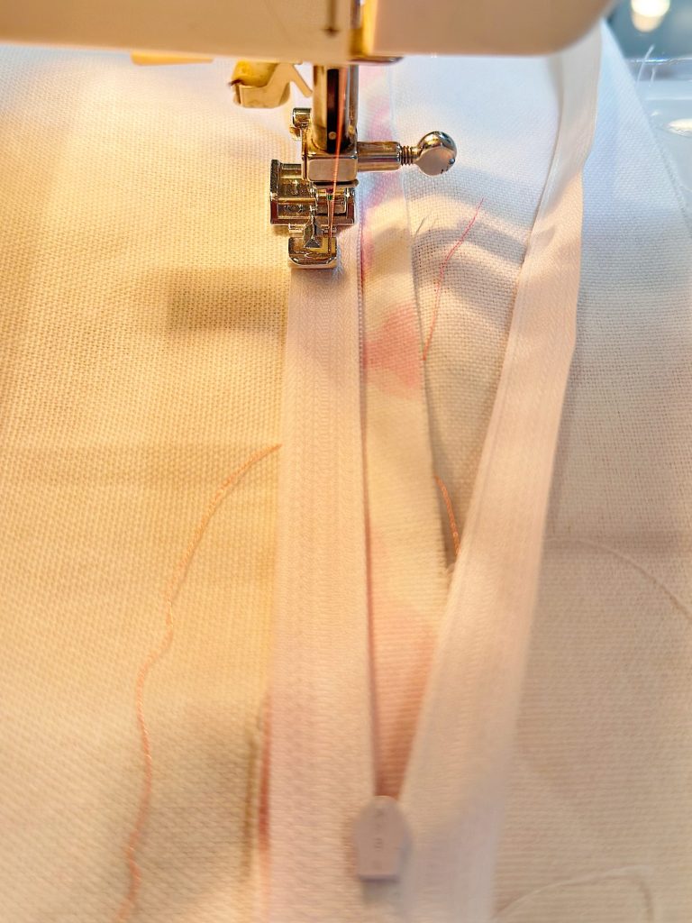 Close-up of a sewing machine stitching a white zipper onto a piece of light-colored fabric.