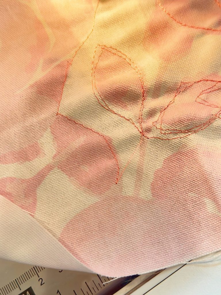 A close-up of pink and white fabric with leaf outlines stitched in red thread; a ruler is partially visible at the bottom left.