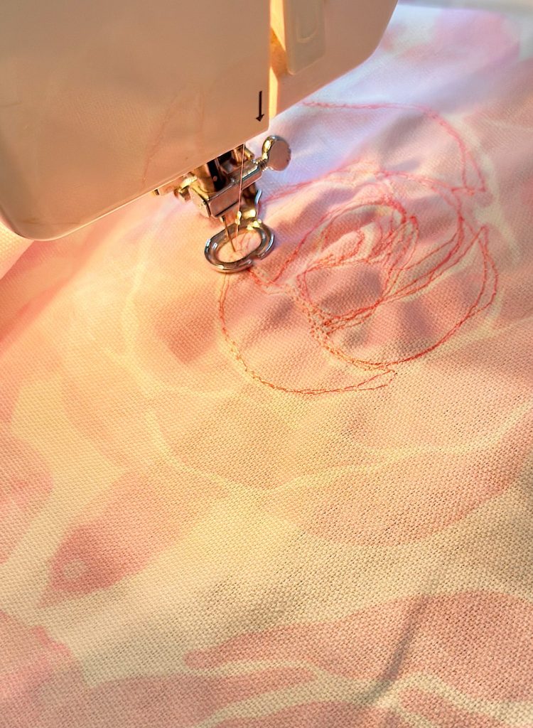 A sewing machine is stitching a rose pattern with pink thread onto light-colored fabric.