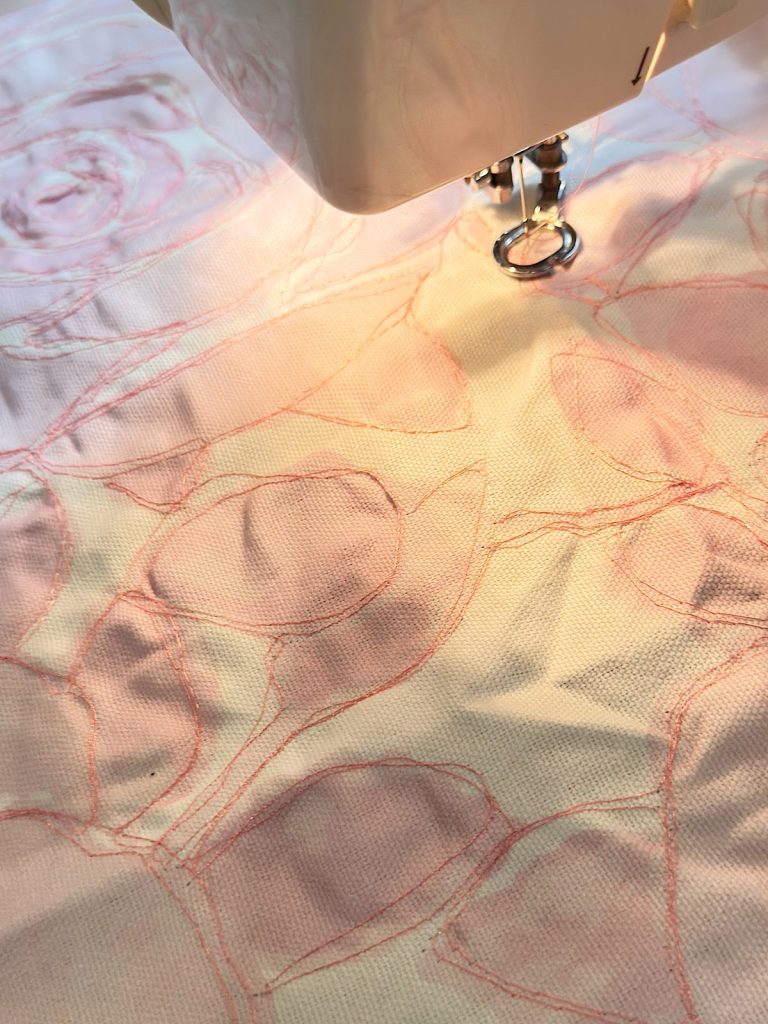 A sewing machine stitching a floral pattern with pink thread on light-colored fabric.