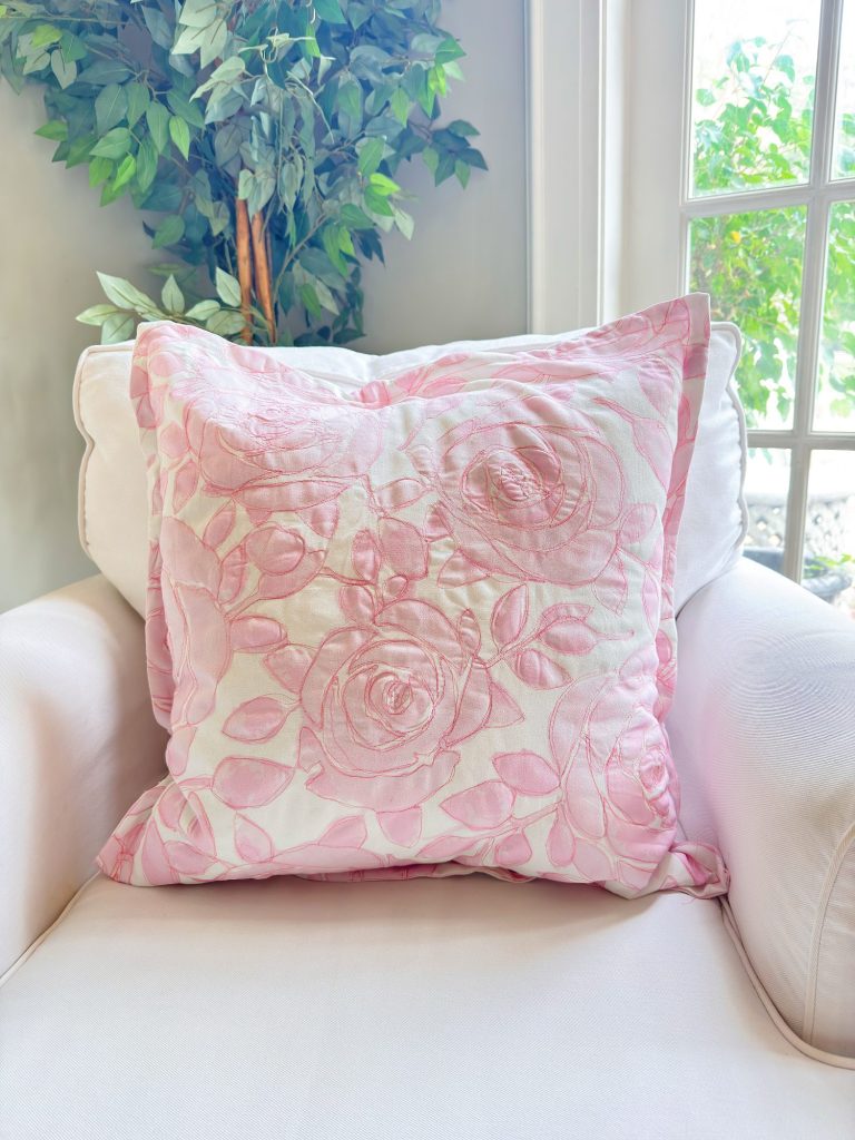 A light pink floral-patterned pillow rests on a white armchair next to a window and a green potted plant.