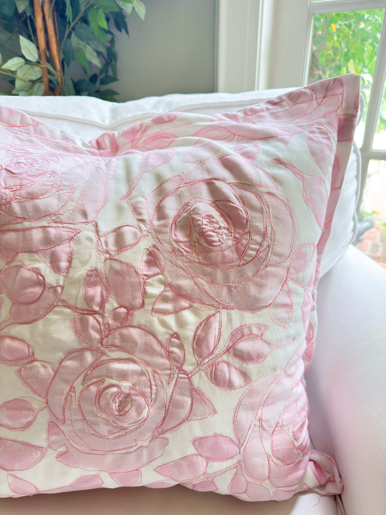 A white pillow with pink embroidered floral designs rests on a white chair near a window with natural light.