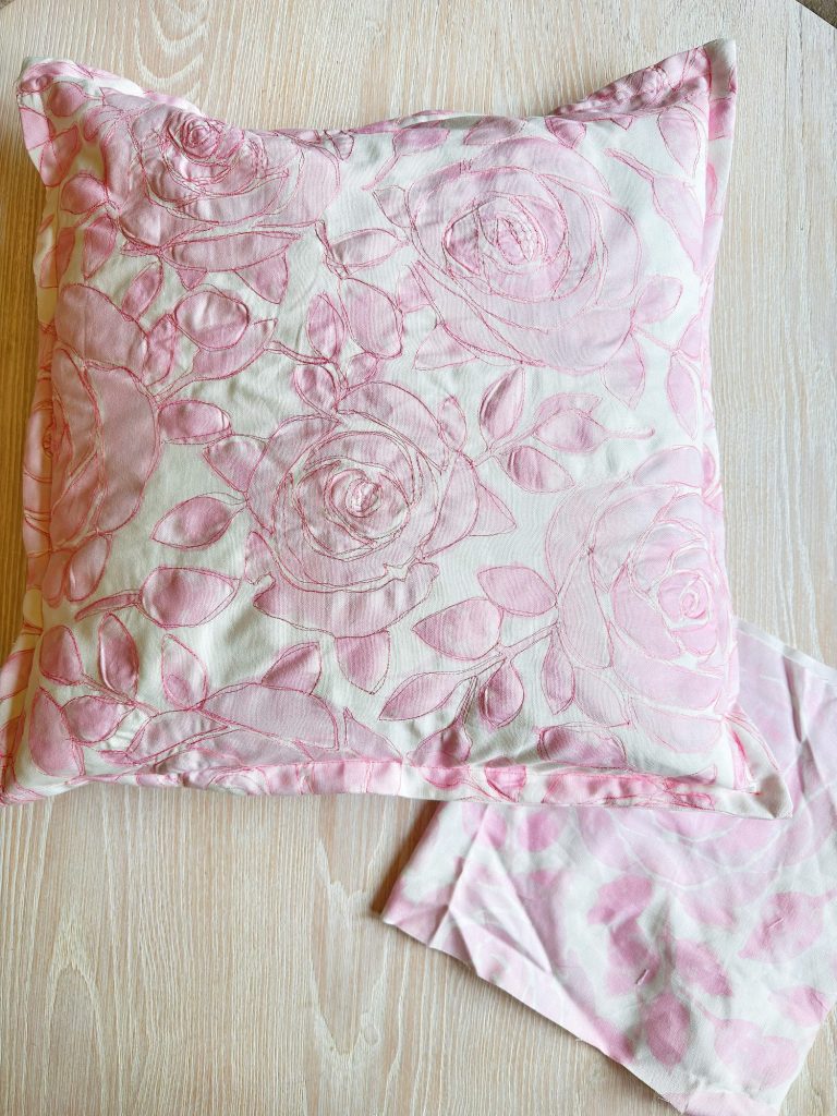 A square pillow with a pink floral pattern and matching fabric rests on a light wood surface.