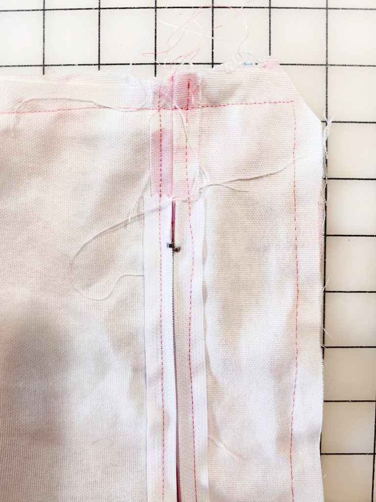 Close-up of a white fabric with a partially sewn zipper and visible pink stitching, lying on a gridded surface.