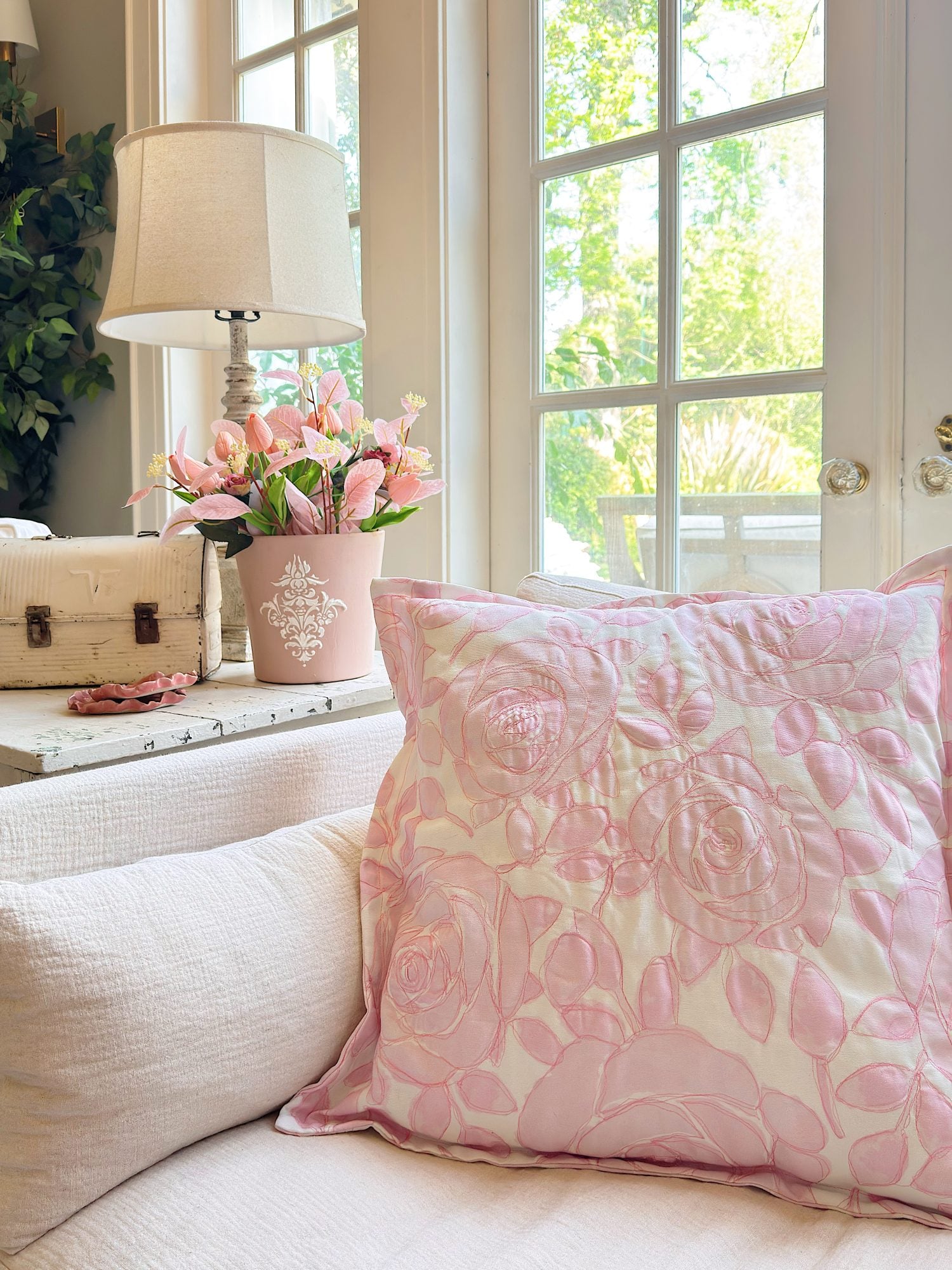 A pink floral pillow on a white couch, with a pink flower arrangement, a white lamp, and a decorative box on a table in front of large windows letting in daylight.