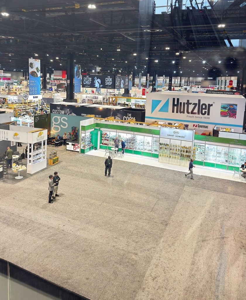 Trade show floor with various branded booths, including Hutzler and Green Sprouts, and a few attendees walking or standing in the open carpeted area.