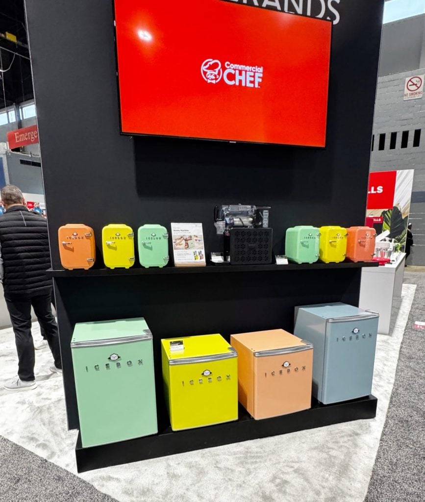 A display booth showcasing colorful Commercial Chef mini fridges and lunchboxes, with a red monitor above and informational materials on the shelf.