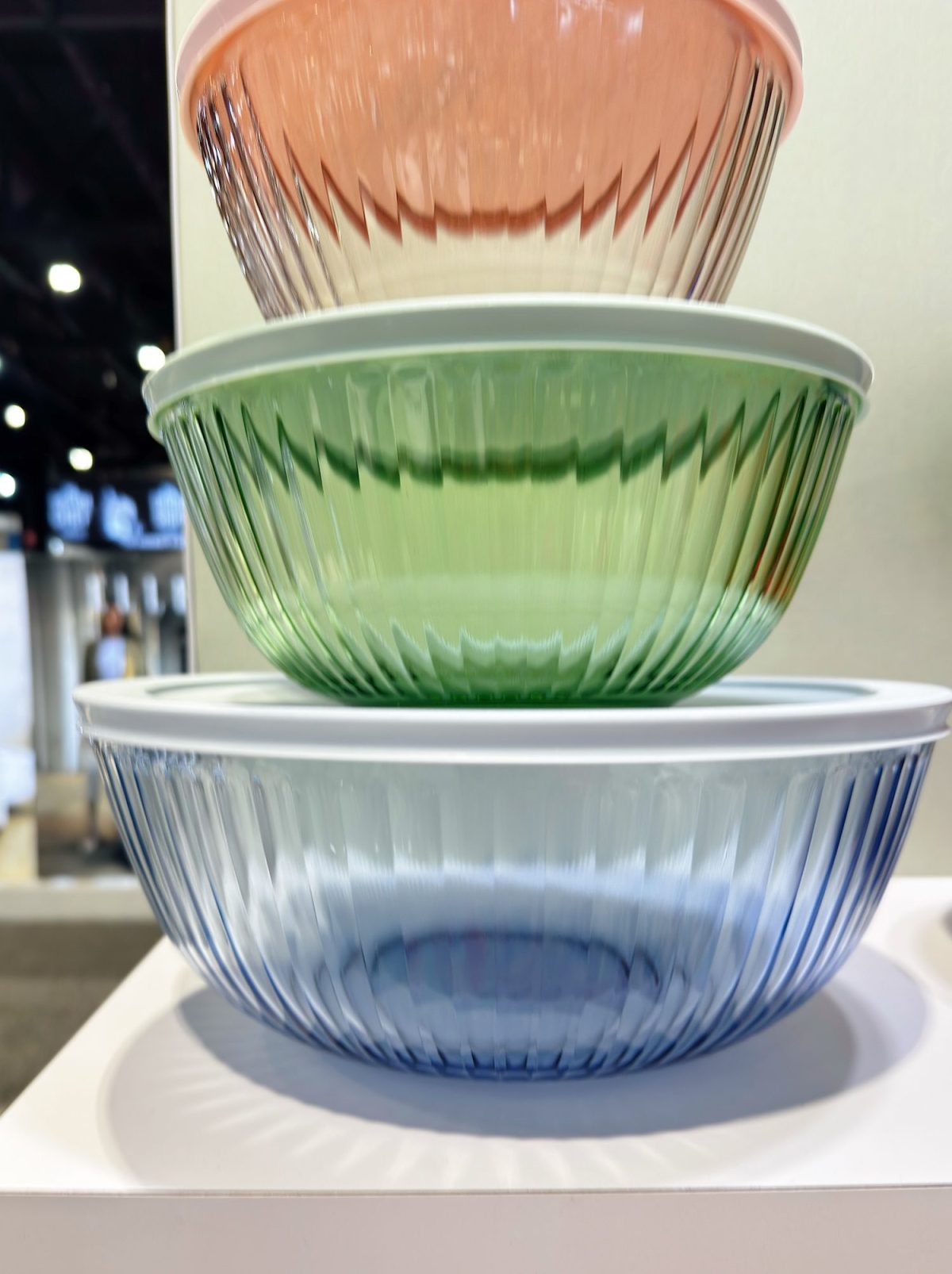 Three ribbed glass bowls in pink, green, and blue, stacked on top of each other, each with a white lid, displayed indoors on a white surface.