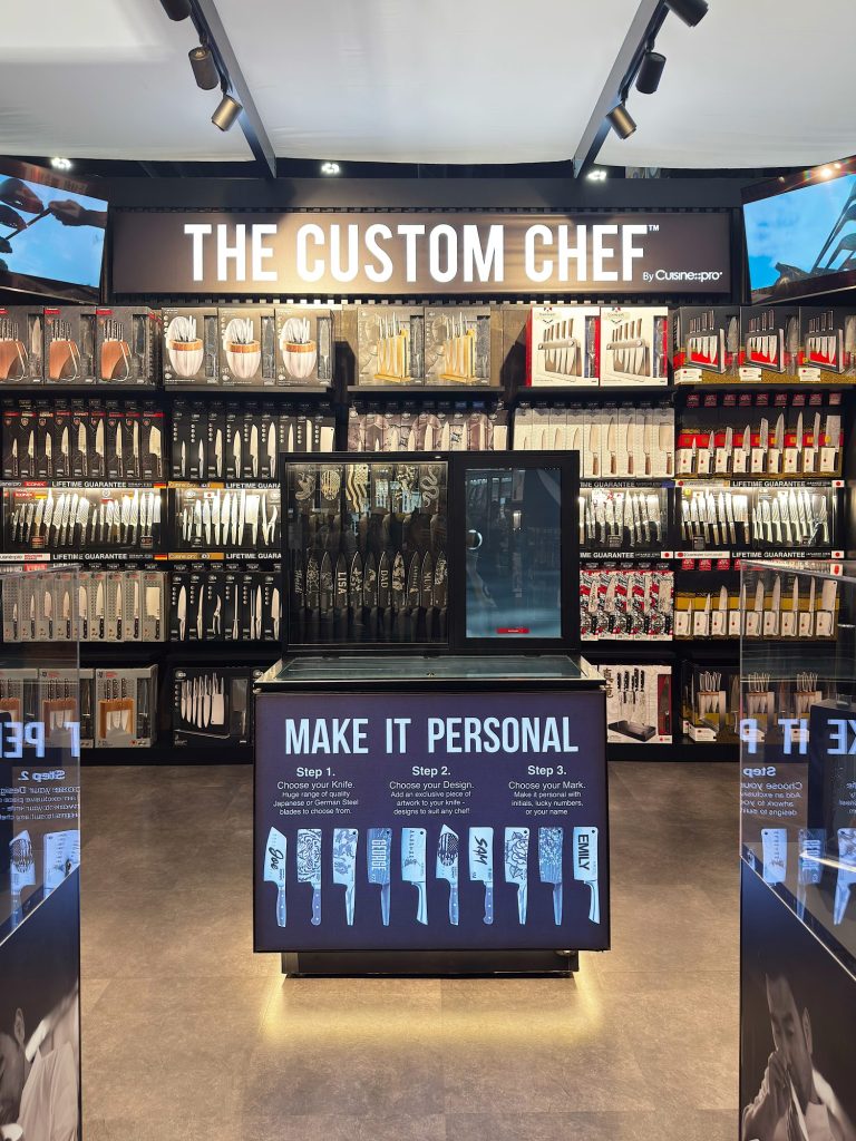 A retail display for "The Custom Chef" showcases various knives on wall shelves, with a central counter offering personalized engraving services for knives.